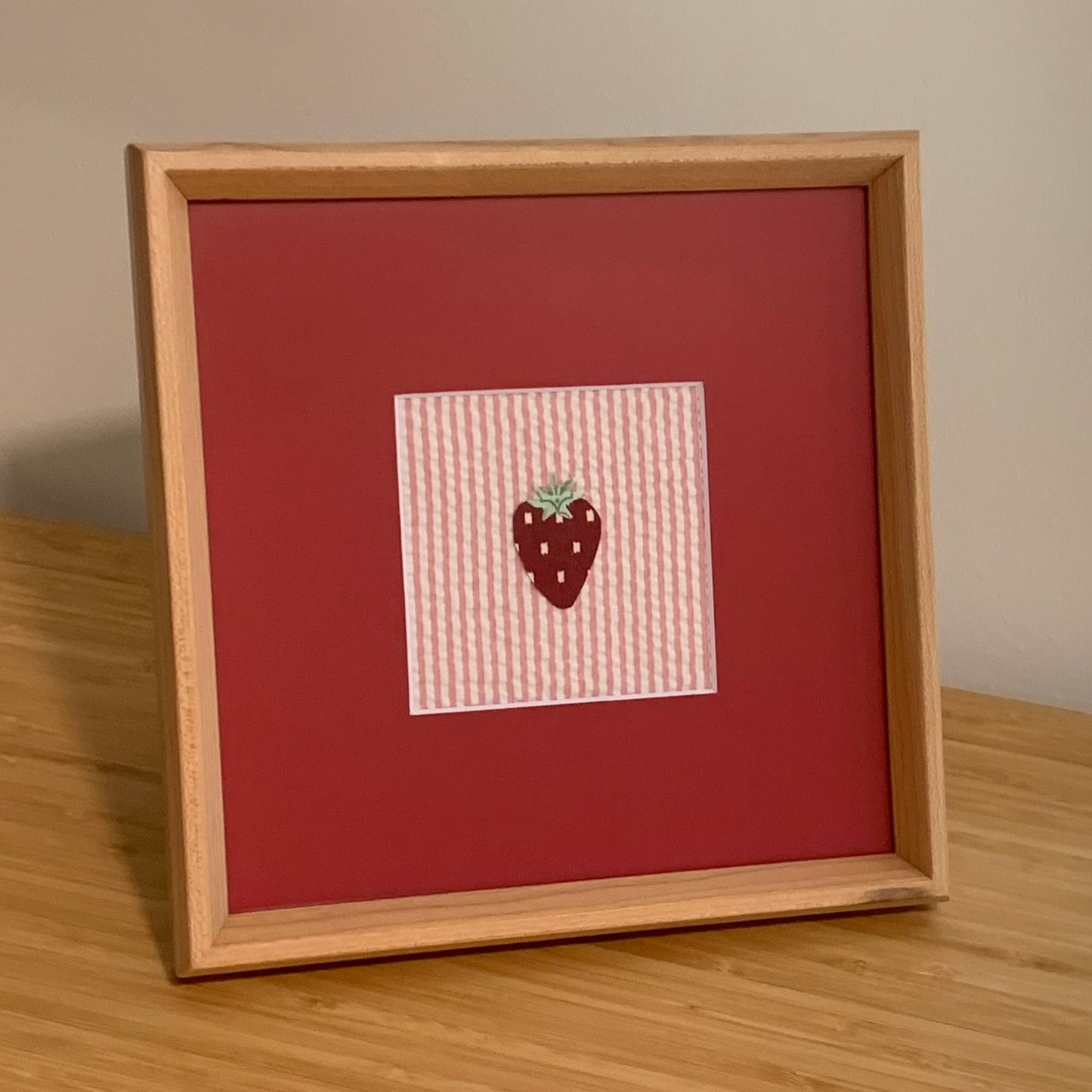 Wooden framed textile artwork of a strawberry on pink and white striped fabric in a red photo mat.