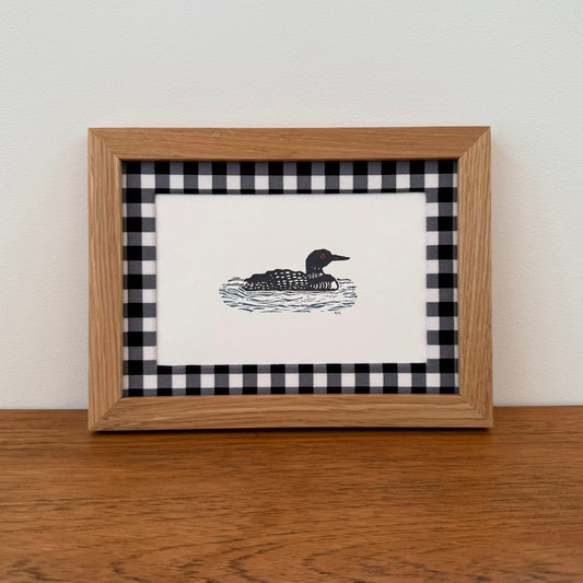 Framed artwork with a loon design on a wooden surface and white wall background