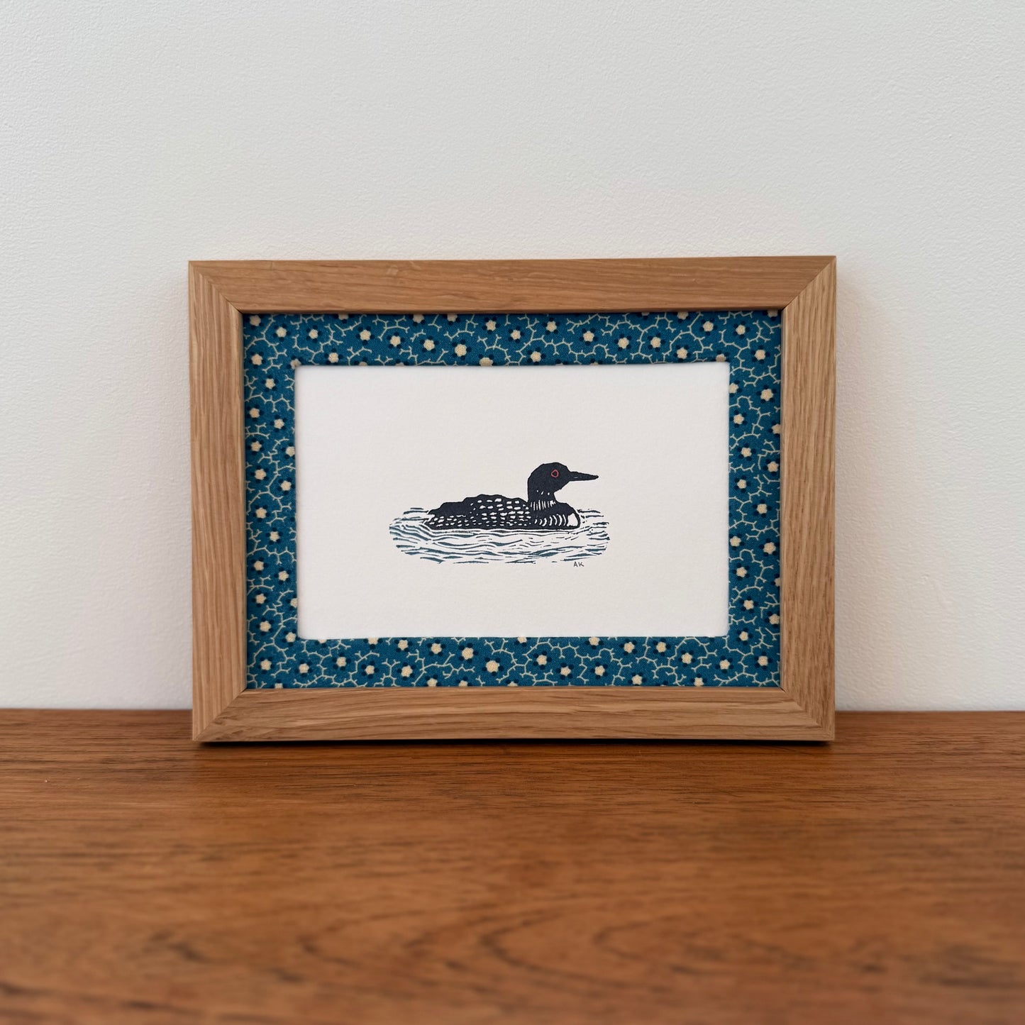 Framed artwork with a loon design on a wooden surface and white wall background