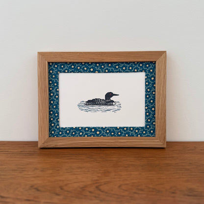 Framed artwork with a loon design on a wooden surface and white wall background