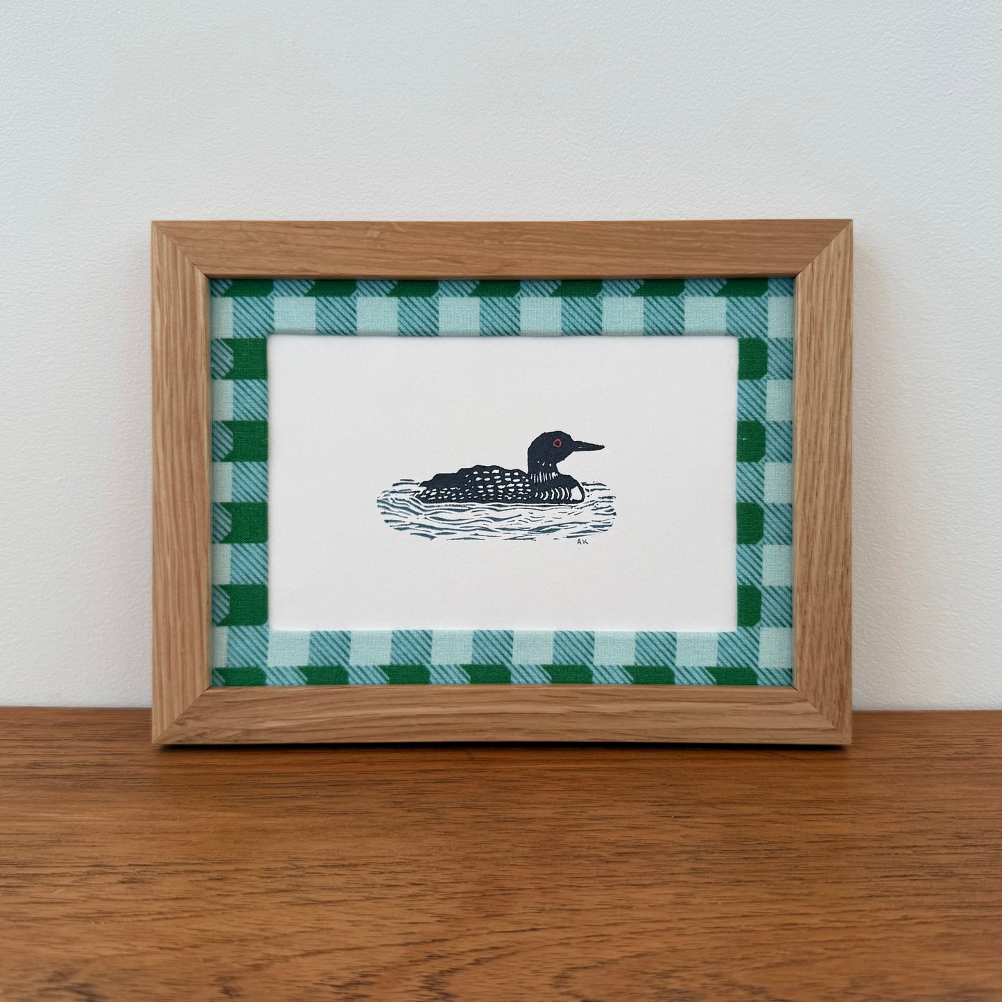 Framed artwork with a loon design on a wooden surface and white wall background
