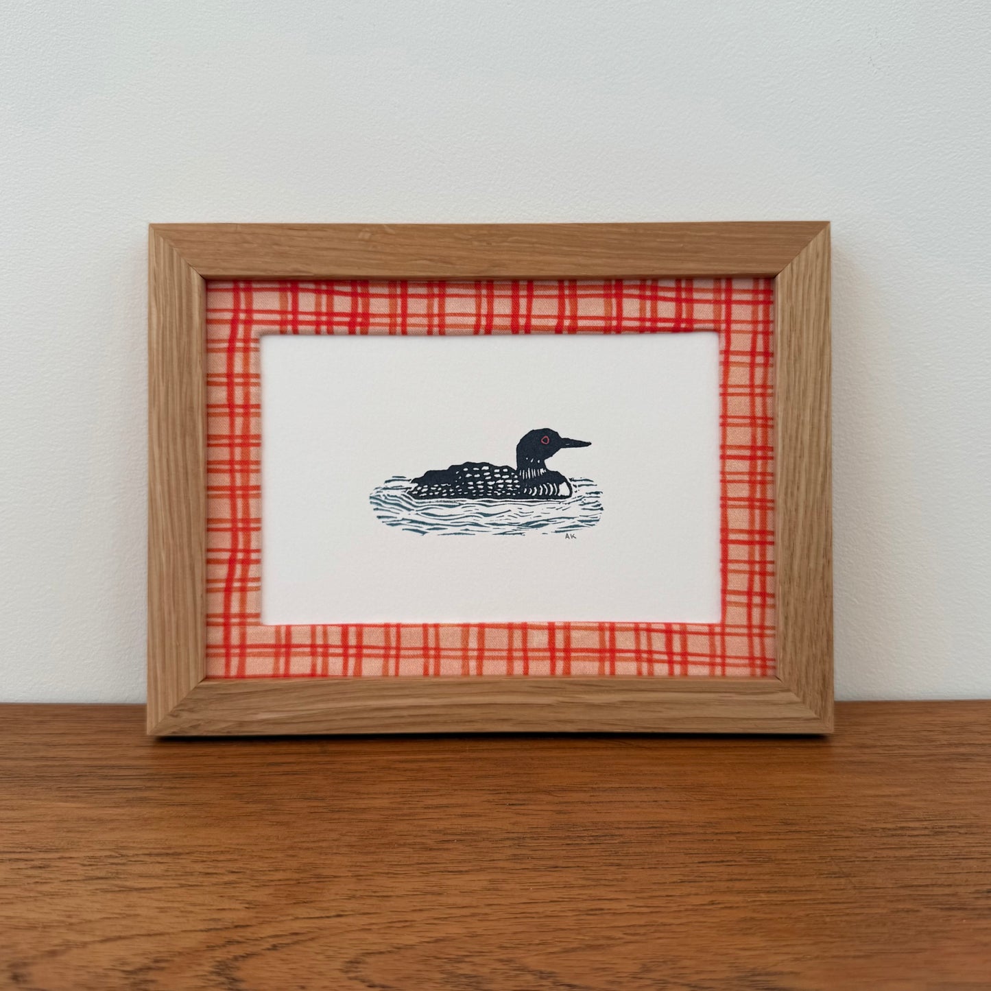 Framed artwork with a loon design on a wooden surface and white wall background