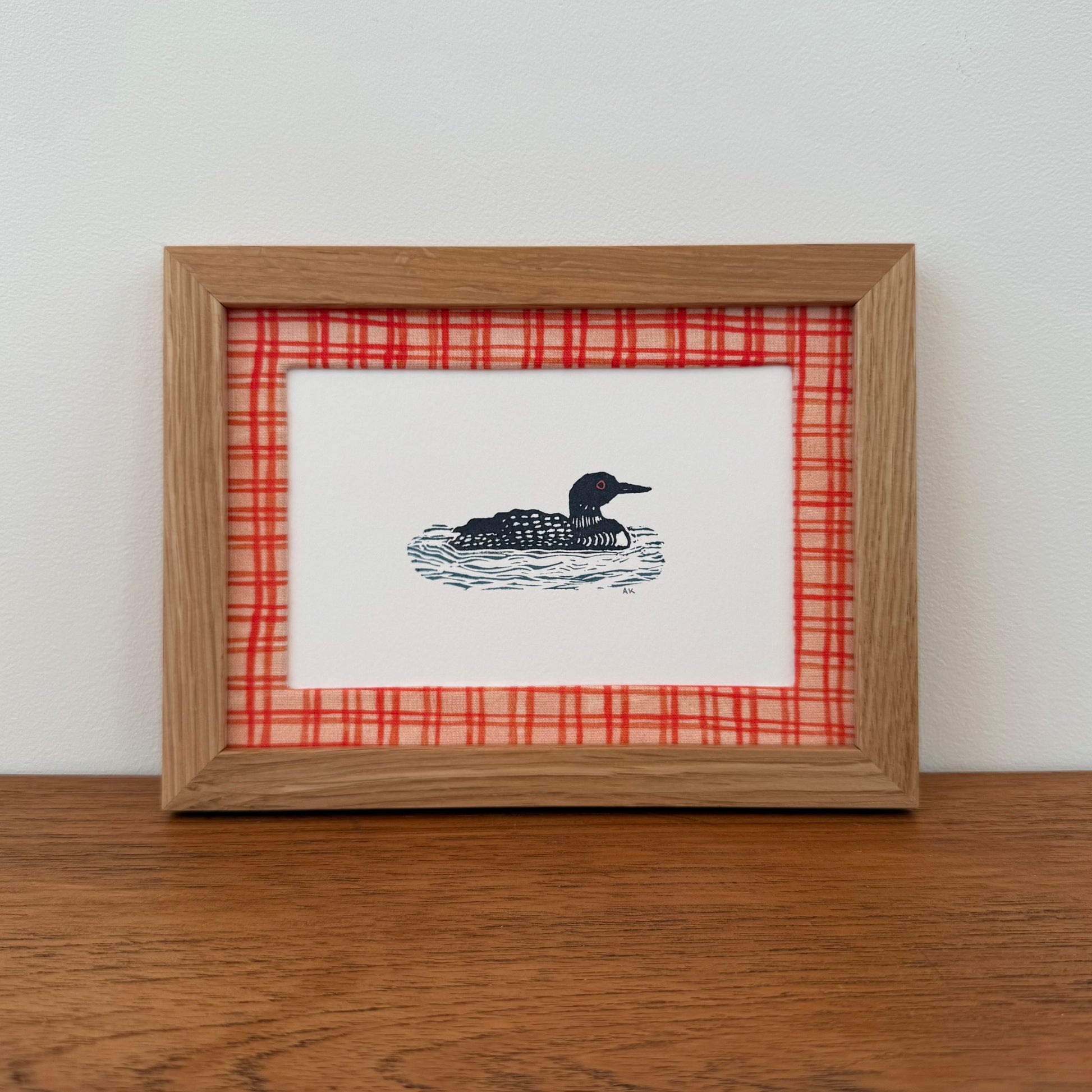 Framed artwork with a loon design on a wooden surface and white wall background