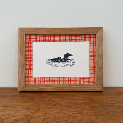 Framed artwork with a loon design on a wooden surface and white wall background