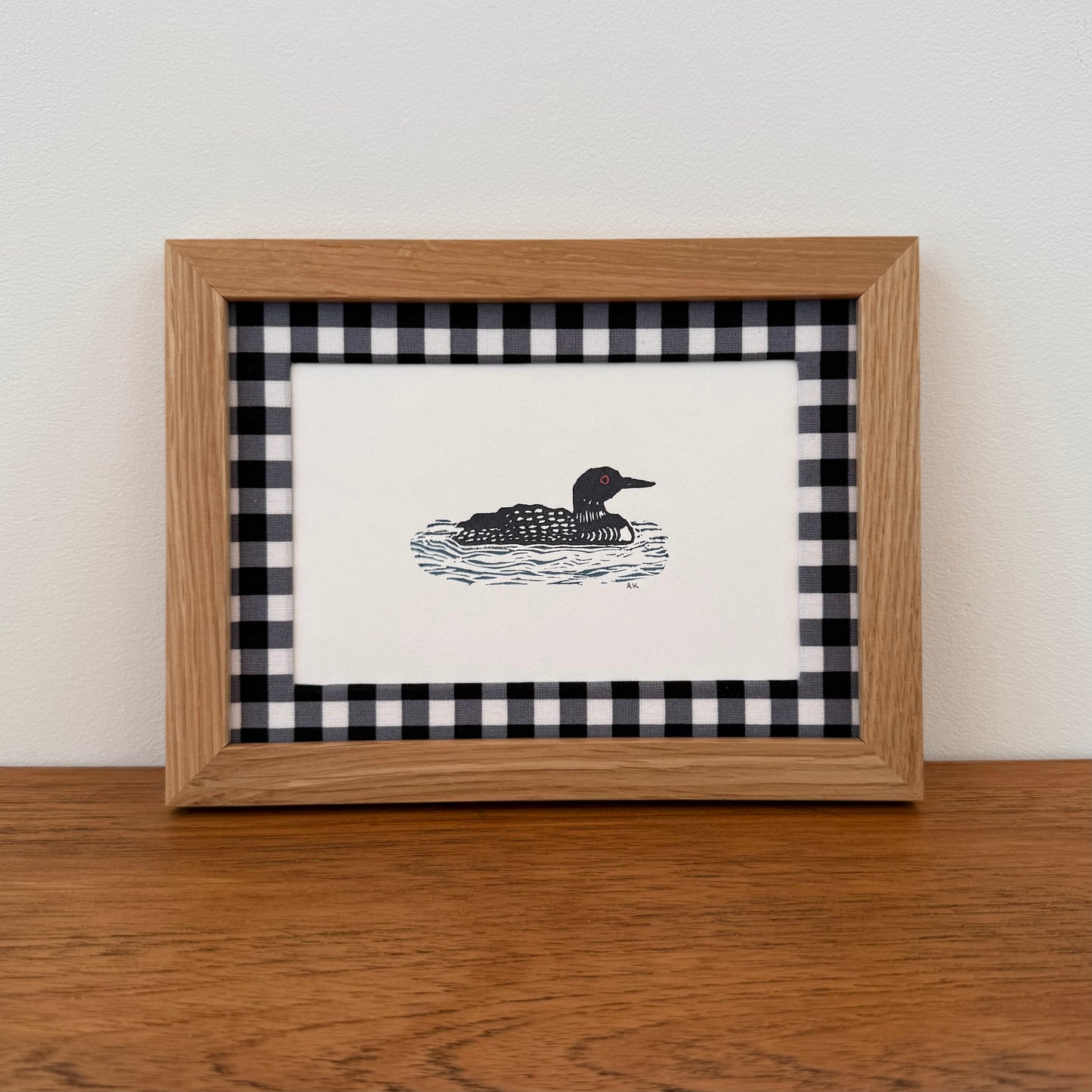 Framed artwork with a loon design on a wooden surface and white wall background