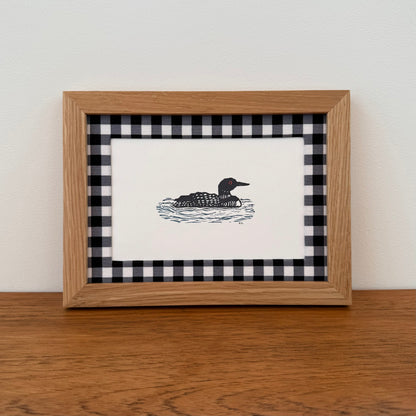 Framed artwork with a loon design on a wooden surface and white wall background