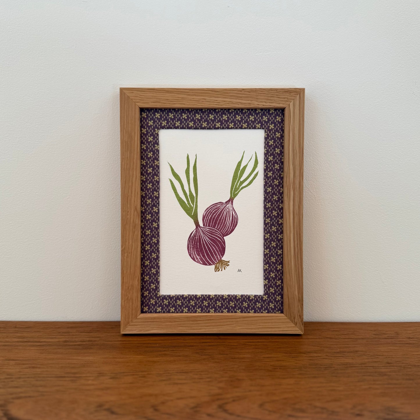 Framed artwork with a red onion design on a wooden surface and white wall background