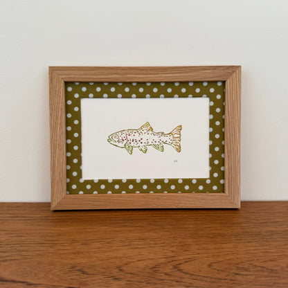 Framed artwork with a trout design on a wooden surface and white wall background