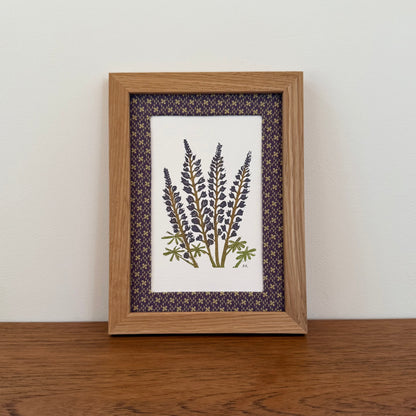 Framed artwork with a lupine design on a wooden surface and white wall background