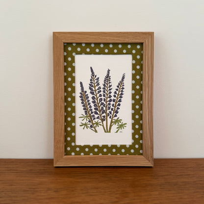 Framed artwork with a lupine design on a wooden surface and white wall background