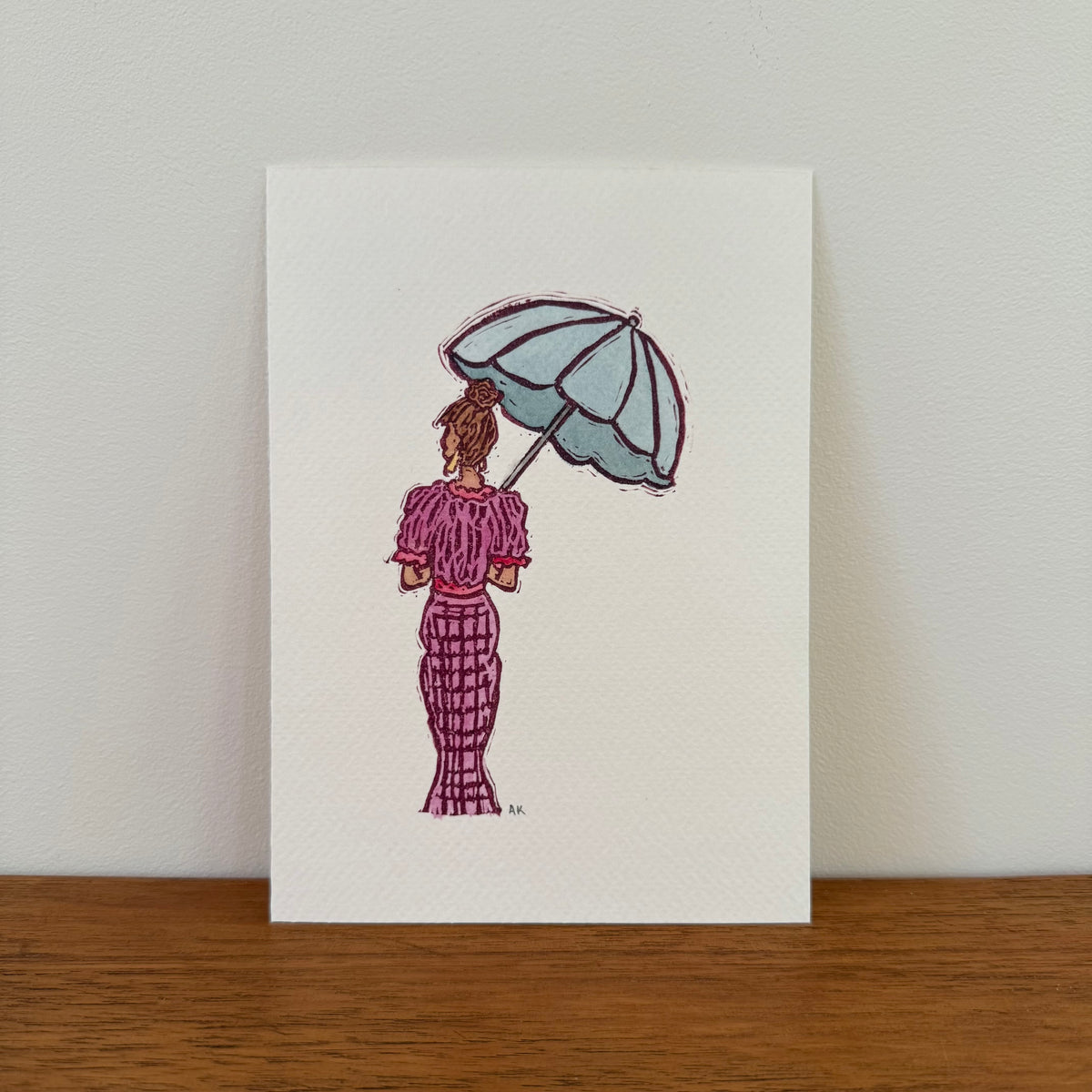 A vertical art print featuring a watercolor illustration of a woman seen from behind, holding a light blue umbrella and wearing a pinkish purple patterned dress, displayed on a wooden surface against a light wall.