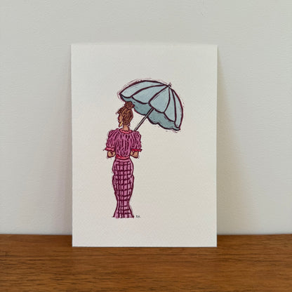 A vertical art print featuring a watercolor illustration of a woman seen from behind, holding a light blue umbrella and wearing a pinkish purple patterned dress, displayed on a wooden surface against a light wall.
