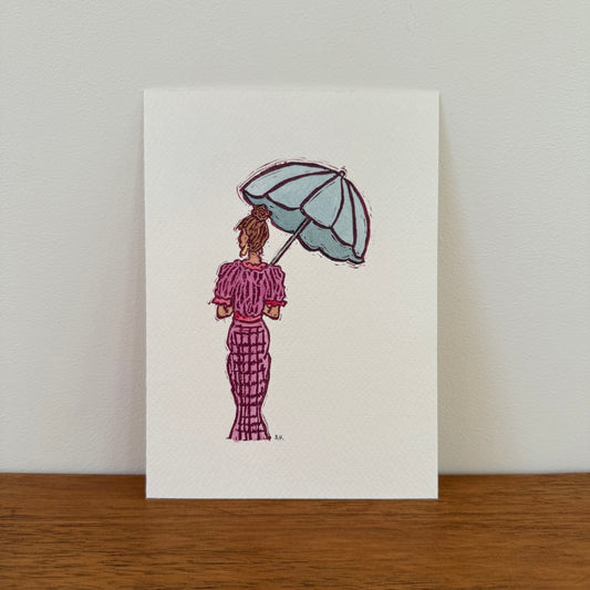 A vertical art print featuring a watercolor illustration of a woman seen from behind, holding a light blue umbrella and wearing a pinkish purple patterned dress, displayed on a wooden surface against a light wall.