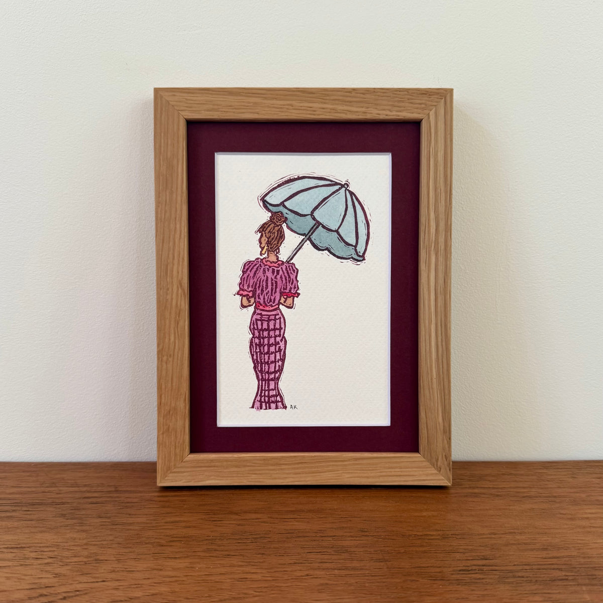 A framed art print featuring a watercolor illustration of a woman seen from behind holding a light blue umbrella, wearing a pinkish purple patterned dress. The print is matted with a deep burgundy mat and housed in a light oak wood frame, displayed on a wooden surface against a white wall.