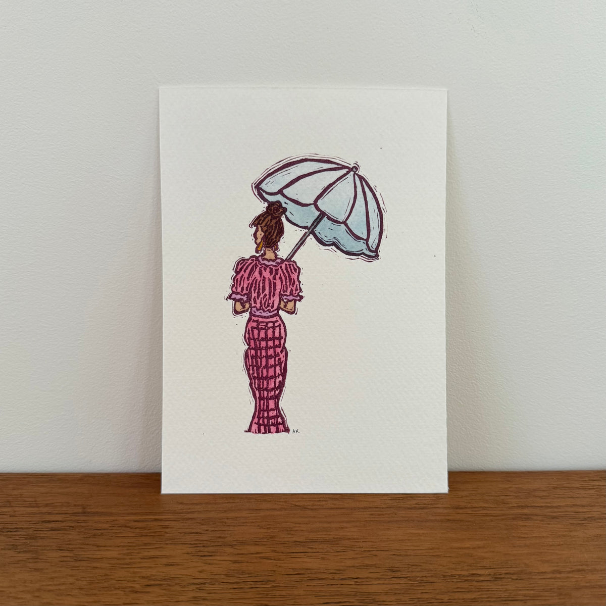A vertical art print featuring a hand-stamped illustration of a woman from behind holding a light blue umbrella, wearing a pink patterned dress. The print is displayed standing upright on a wooden surface against a plain off-white wall.