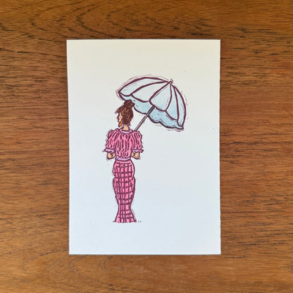 A vertical art print featuring an illustration of a woman seen from behind holding a light blue and white umbrella, wearing a pink patterned dress. The print is displayed flat on a brown wooden surface.