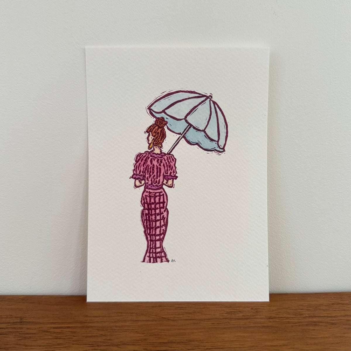 A vertical art print featuring a hand-stamped illustration of a woman seen from behind holding a light blue umbrella, wearing a textured magenta dress. The print is displayed standing upright on a wooden surface against a neutral off-white wall.