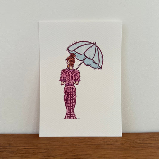 A vertical art print featuring a hand-stamped illustration of a woman seen from behind holding a light blue umbrella, wearing a textured magenta dress. The print is displayed standing upright on a wooden surface against a neutral off-white wall.
