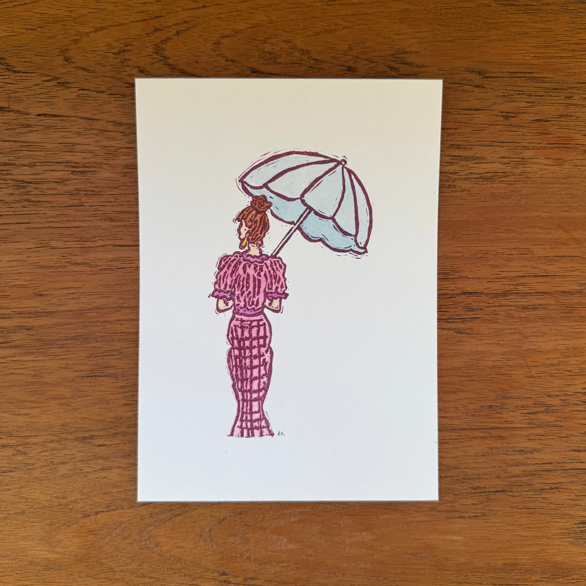 A vertical art print featuring a hand-stamped illustration of a woman in a pink dress holding a light blue umbrella, placed on a wooden tabletop.