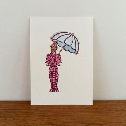A vertical art print featuring a hand-stamped illustration of a woman seen from behind, holding a light blue umbrella and wearing a pink patterned dress, displayed on a wooden surface against a light wall.