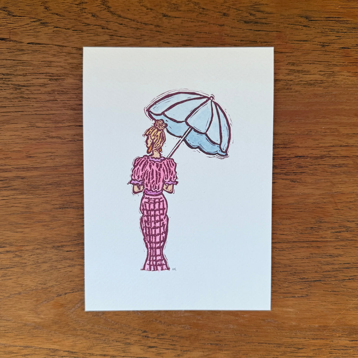 An art print featuring a hand-stamped illustration of a woman in a pink dress holding a light blue umbrella, displayed flat on a brown wooden surface.
