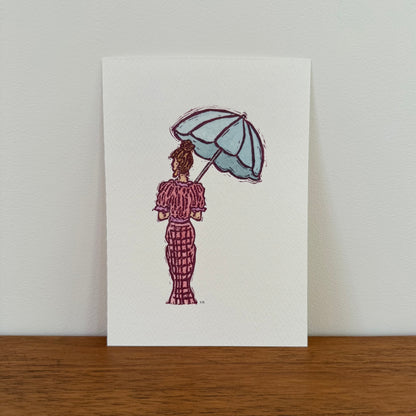 A vertical art print featuring a hand-stamped watercolor illustration of a woman from behind holding a light blue umbrella, standing on a wooden surface against a light gray wall.