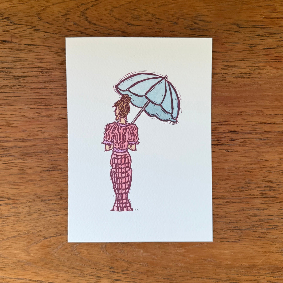 Art print featuring a hand-stamped illustration with watercolor of a woman from behind holding a light blue umbrella, placed on a brown wooden surface.
