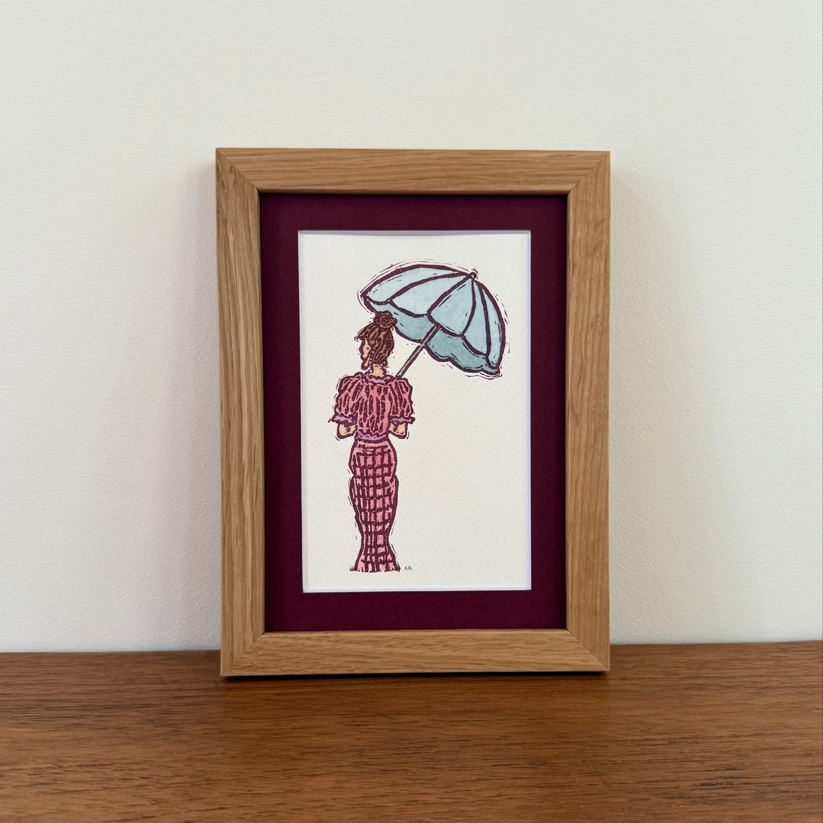 A framed art print featuring a hand-stamped illustration with watercolor of a woman from behind holding a light blue umbrella, set in a light oak frame with a deep burgundy mat, displayed on a wooden surface against a white wall.