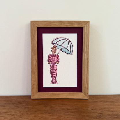 A framed art print featuring a hand-stamped illustration of a woman in a pink patterned dress holding a light blue umbrella, set against a white background with a deep burgundy mat and a light oak wood frame, displayed on a wooden surface against a white wall.