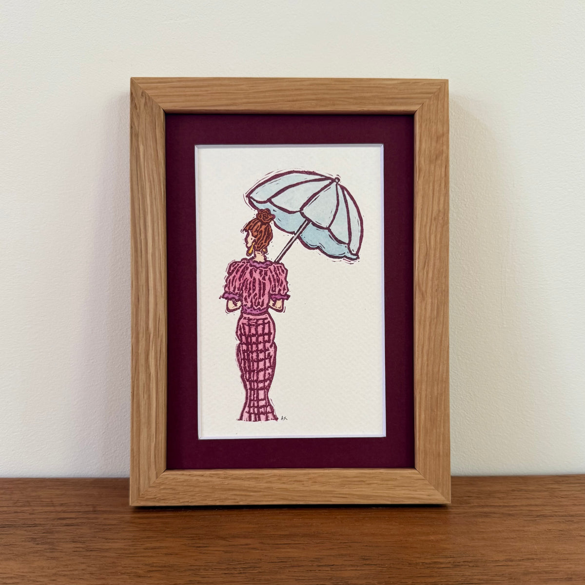 A framed vertical art print with a burgundy photo mat,  featuring a hand-stamped illustration of a woman in a pink dress holding a light blue umbrella, placed on a wooden tabletop.