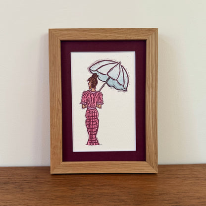 A framed art print featuring a hand-stamped illustration of a woman in a pink patterned dress holding a light blue umbrella, set in a light oak frame with a dark burgundy mat on a wooden table against a white wall.