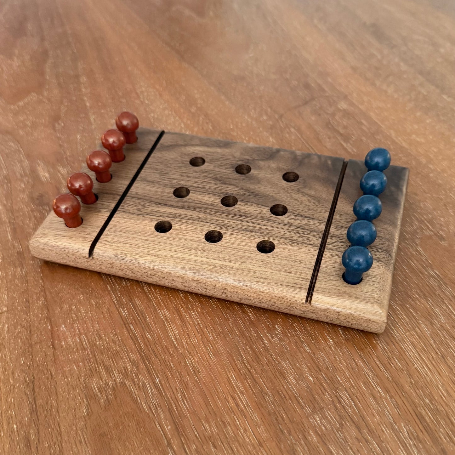 a wooden tic-tac-toe game board with rust and blue colored playing pegs, sitting on a wooden table.