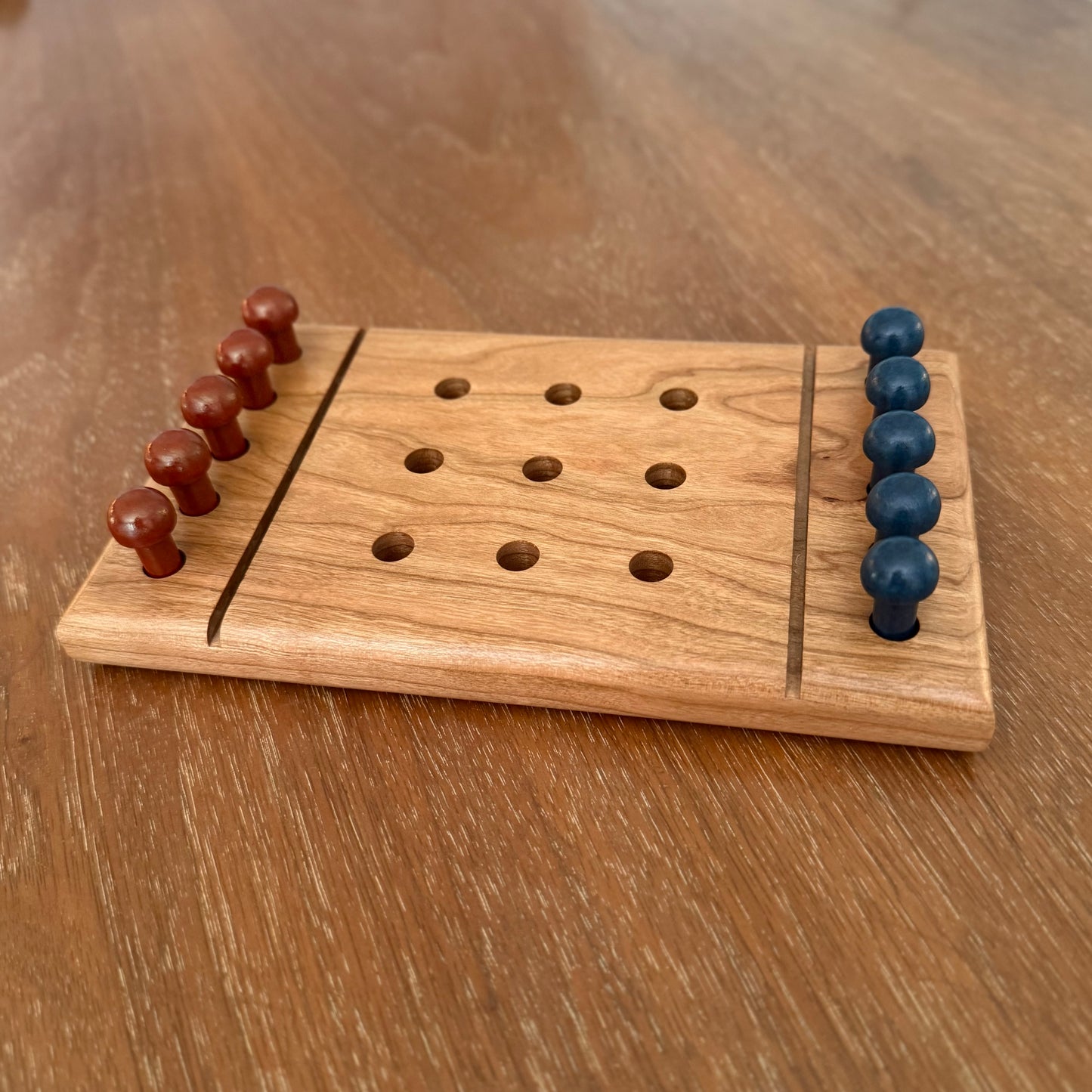 A small wooden tic-tac-toe board game with carved holes and two rows of round wooden playing pieces, one set in reddish brown and one set in dark blue, placed on a wooden tabletop.