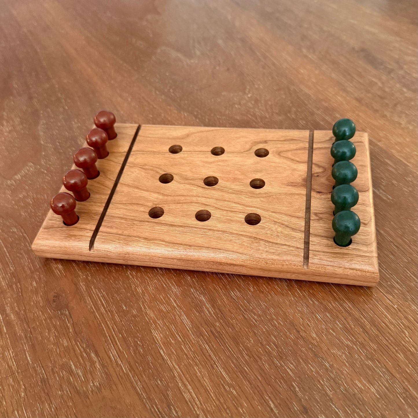 A small wooden tic-tac-toe board game with carved holes and two rows of round wooden playing pieces, one set in reddish brown and one set in dark green, placed on a wooden tabletop.