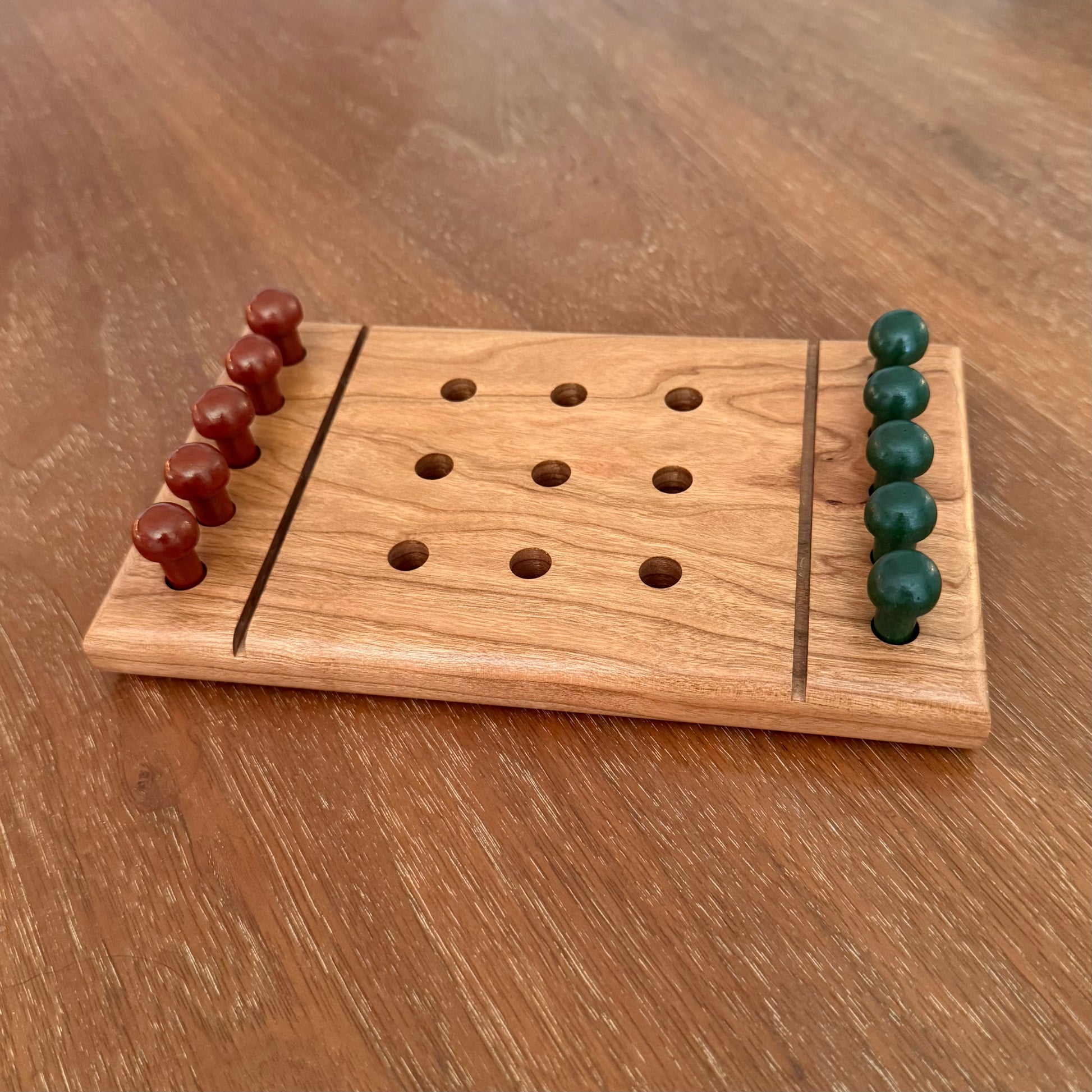 A small wooden tic-tac-toe board game with carved holes and two rows of round wooden playing pieces, one set in reddish brown and one set in dark green, placed on a wooden tabletop.