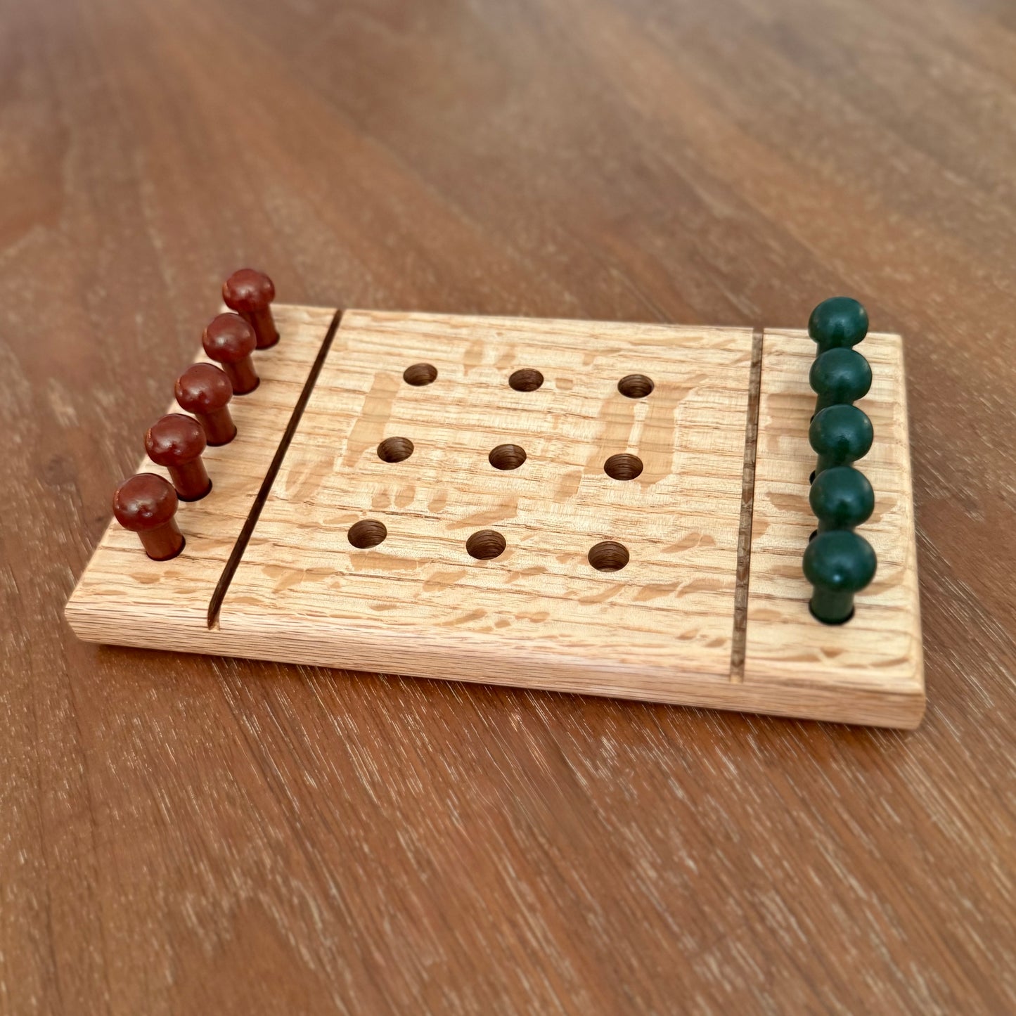 A small wooden tic-tac-toe game board  sitting on a table with rust colored playing pegs and dark green colored playing pegs.