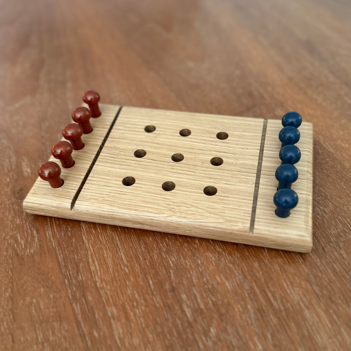 A small wooden white oak tic-tac-toe game board set on a wooden table with reddish brown playing pegs and blue playing pegs.