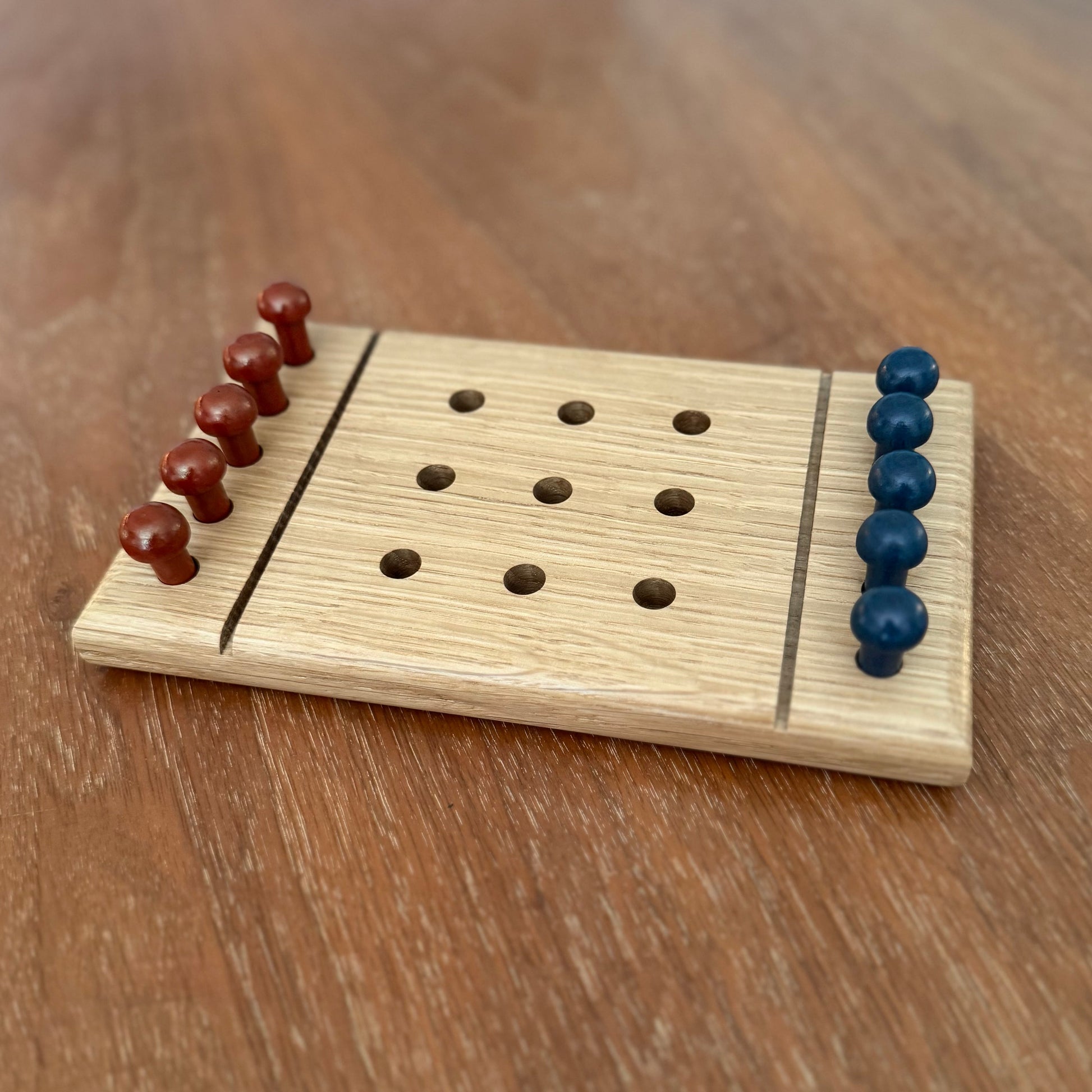 A small wooden white oak tic-tac-toe game board set on a wooden table with reddish brown playing pegs and blue playing pegs.