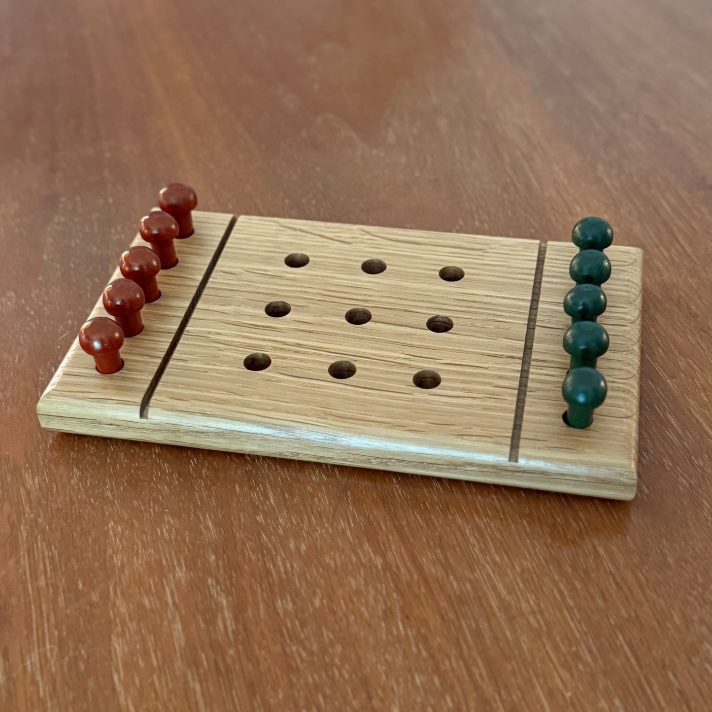 A small wooden white oak tic-tac-toe game board set on a wooden table with reddish brown playing pegs and green playing pegs.